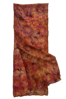 A lightweight scarf with an all-over floral or sunburst batik pattern in warm autumn colors, including rust orange, terracotta red, and brown.