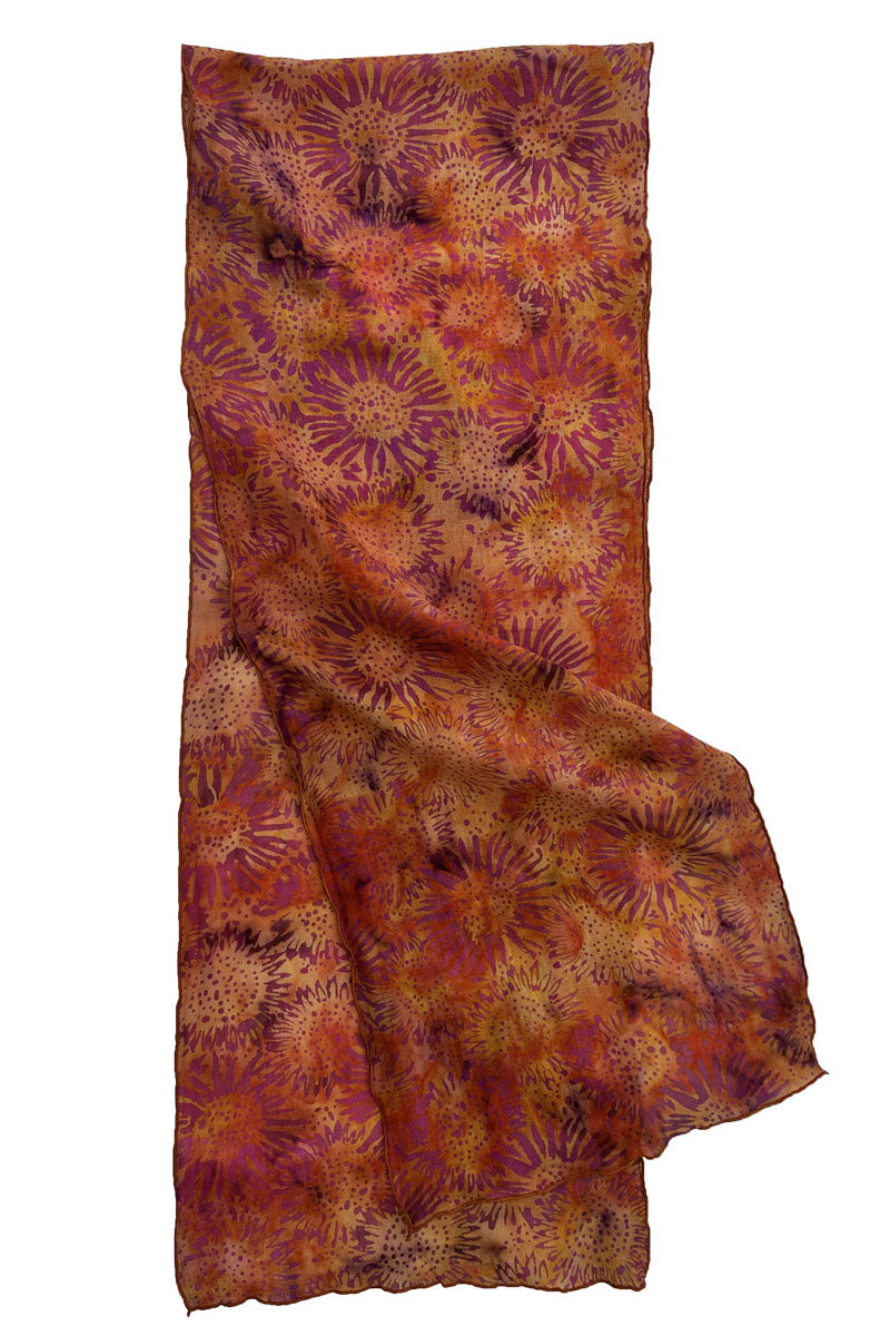 A lightweight scarf with an all-over floral or sunburst batik pattern in warm autumn colors, including rust orange, terracotta red, and brown.