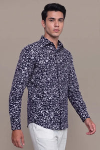 A man wearing a navy blue long-sleeve button-up shirt with a white floral pattern, paired with white trousers, standing against a solid beige background.
